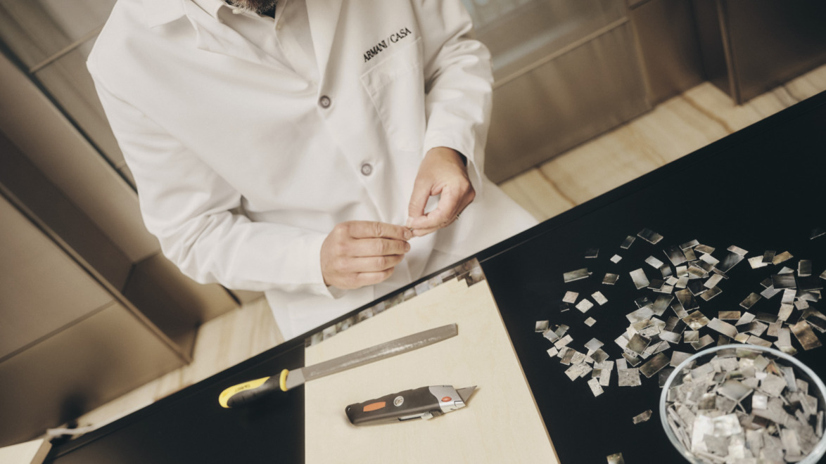 A person in a white lab coat carefully arranging small tiles on a black table with a utility knife nearby.